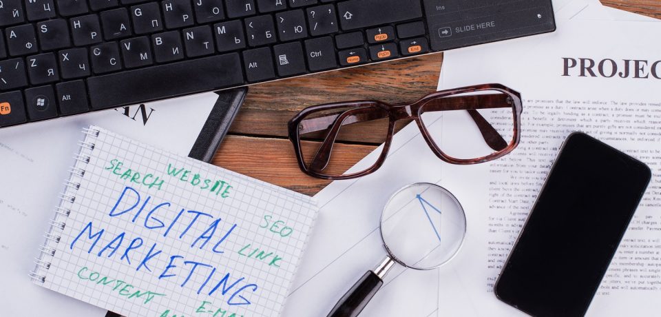 Digital marketing on notepad and various business papers on brown background. Brown glasses magnifier and smartphone near notepad.