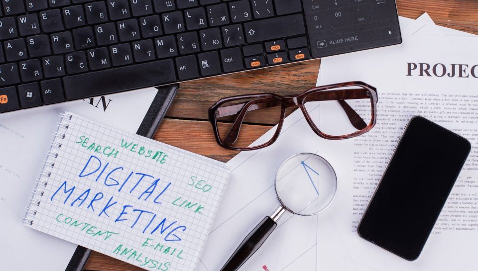 Digital marketing on notepad and various business papers on brown background. Brown glasses magnifier and smartphone near notepad.
